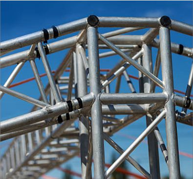 A close-up architectural shot of silver aluminum circular stage trussing connectors against a clear blue sky, showing professional event rigging equipment.