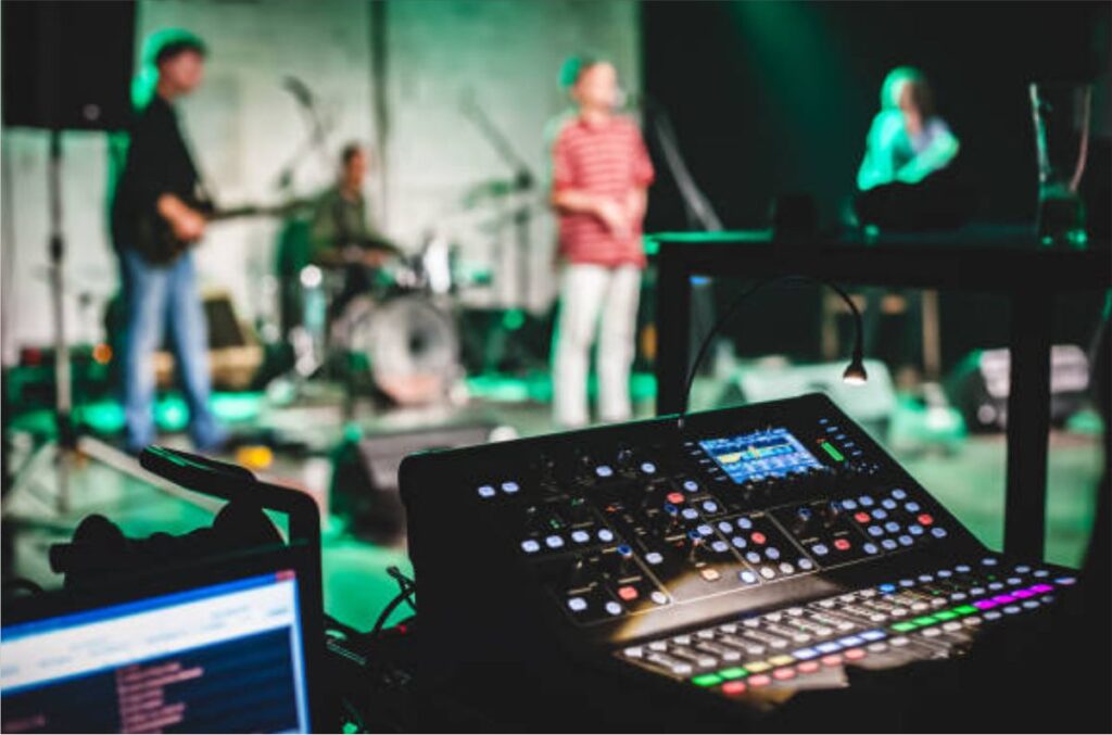 Close-up of a digital audio mixing console with glowing buttons and faders, with a live band performing on a lit stage in the blurred background.