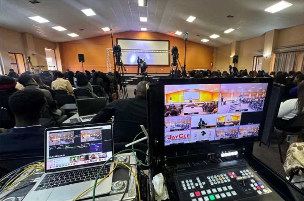 An expansive view of a live event production desk equipped with a laptop, video switcher, and multi-view monitor overlooking a crowded hall with a speaker on stage.
