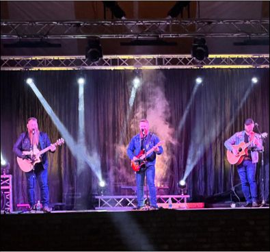Three-piece band performing on a stage with vertical light beams and atmospheric smoke effects.