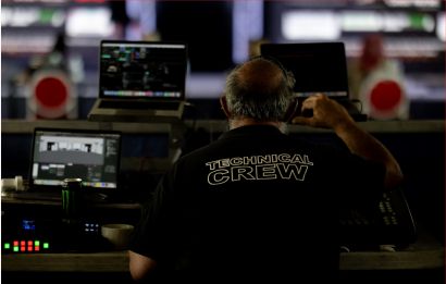 A technical crew member in a black shirt monitors multiple laptop screens and a broadcast console during a live event production.