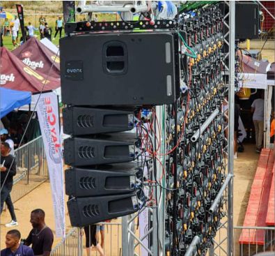 A vertical line array of professional event speakers mounted on a metal truss at an outdoor festival or sporting event.