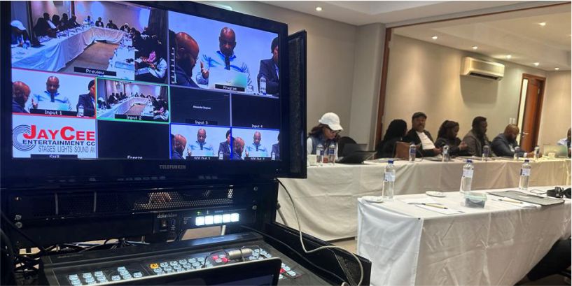 A professional live streaming control station featuring a multi-view monitor showing four camera inputs, a hardware video switcher, and an event production crew in the background.